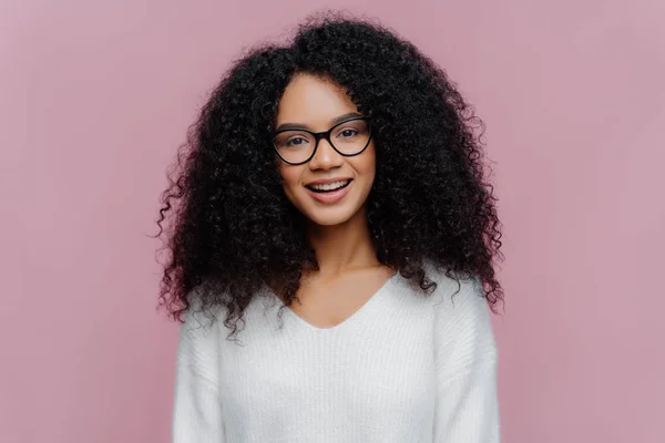 Портрет приємної усміхненої жінки з африканською зачіскою, в оптичних окулярах і білому светрі, має веселий вираз обличчя, ізольований над фіолетовим фоном. Тисячолітня дівчинка постає у приміщенні — стокове фото