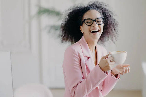 Mujer de pelo rizado muy alegre se ríe felizmente mientras bebe café caliente o café con leche, lleva gafas transparentes, se divierte durante el descanso en la oficina, muestra los dientes blancos, siendo empresario profesional —  Fotos de Stock