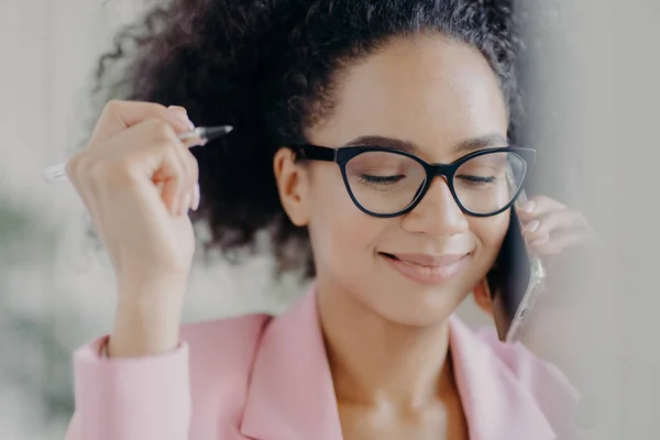 Headshot of delighted female director holds pen in hand, wears optical eyeglasses, has gentle smile, makes call via modern cell phone, dressed in pink elegant costume, focused down with shyness