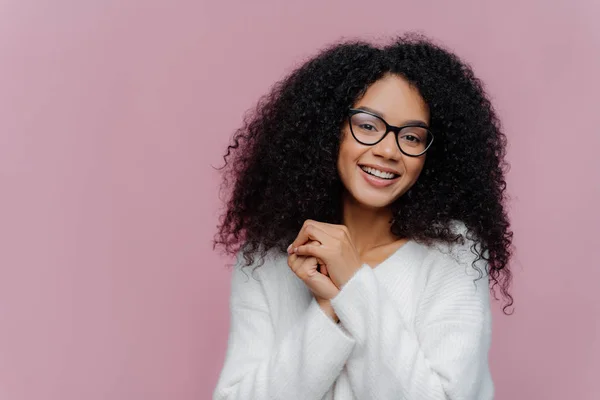 Portrait de heureuse femme afro-américaine positive aux cheveux bouclés et touffus, garde les mains jointes, a une expression faciale agréable, porte des lunettes et un pull blanc, pose sur fond violet — Photo