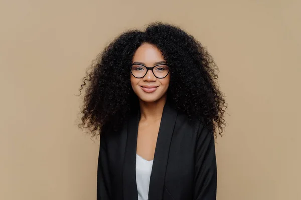 Porträtt av vacker afro amerikansk kvinna med krispigt hår, klädd i elegant svart jacka, transparent glasögon, tittar direkt på kameran med milt leende bär optiska glasögon isolerade på brun vägg — Stockfoto