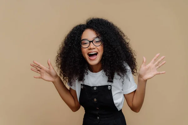 Överlycklig afroamerikansk kvinna skrattar ut, höjer handflatorna, är i hög anda, bär genomskinliga glasögon, casual t skjorta och overaller, poserar mot brun bakgrund, känner lycka och nöje — Stockfoto