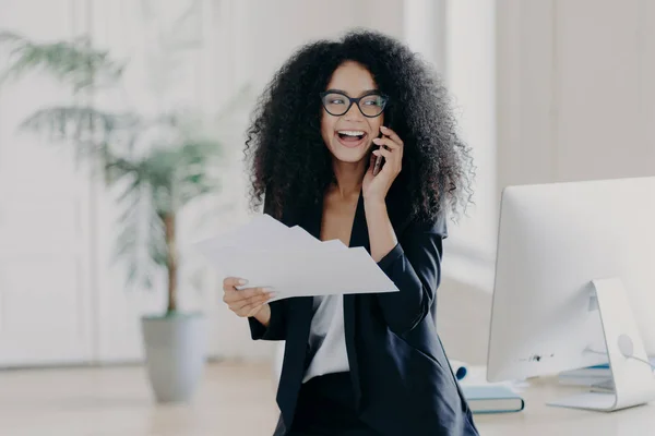 Pleased curly haired female director dressed in formal outfit, holds documents, calls via cell phone, stands near workplace with computer, shares opinion about startup, makes business report