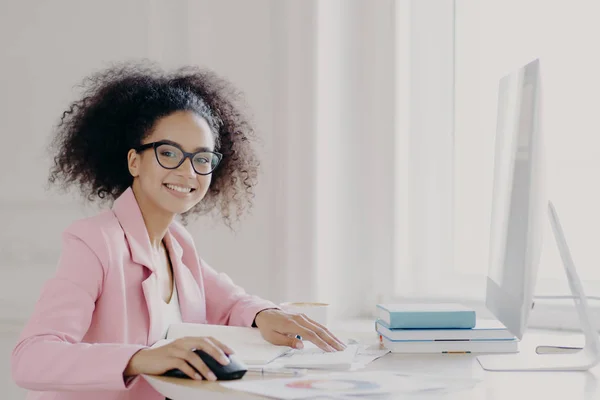 Velstående kvinnelige entreprenør sjekker dokumenter, studiekontrakter, arbeid på datamaskin, bruker briller og rosa jakke, poserer på arbeidsplassen, smiler bredt, er nesten ferdig i tide – stockfoto