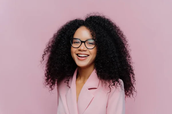 Femme d'affaires bouclée positive porte des lunettes, tenue formelle, a une coiffure touffue, étant de bonne humeur après une journée réussie au travail, pose sur fond violet. Monochrome. Heureux réalisateur — Photo