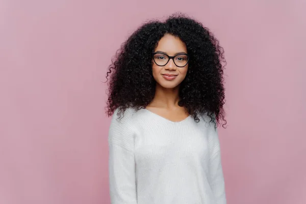Plan à mi-longueur de femme afro-américaine attrayante regarde à travers des lunettes transparentes, pull blanc, a une expression calme grave, pose contre le mur du studio violet. Concept d'expressions faciales — Photo