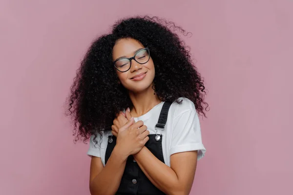 Porträtt av nöjd kvinna med afro frisyr, håller händerna pressade på bröstet, lutar huvudet, bär vit casual t skjorta och overaller, poser över lila bakgrund, minns trevligt ögonblick i livet. — Stockfoto