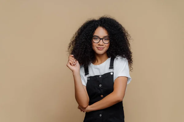 Isolerad skott av trevlig ser ung afroamerikansk kvinna vidrör lockigt hår, har buskig frisyr, bär optiska glasögon, vit t skjorta och svarta overaller, poser över brun studio bakgrund — Stockfoto