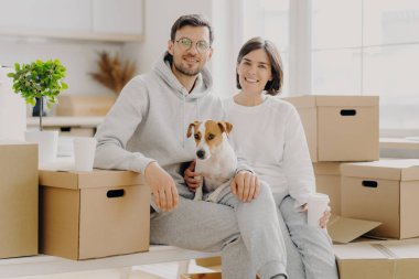 Pozitif kadın ve erkek karton kutuların yanında poz veriyor, köpekle resim yapmak için poz veriyor, yeni bir yere yerleşiyor, beyaz günlük kıyafetler giyiyor, geniş pencereli bir mutfakta poz veriyor.