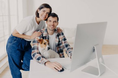 Mutlu aile fotoğrafı bilgisayar monitörünün yanında duruyor, uzaktan birlikte çalışıyorlar, kendi işleri var, internetten para kazanıyorlar, kameraya zevkle bakıyorlar, favori evcil hayvanla oynuyorlar. İnsanlar, teknoloji.