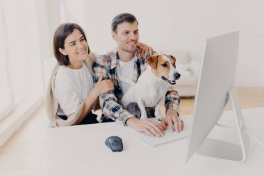 Neşeli karı ve kocaların kapalı çekimleri neşeli ifadelerle bakın, komik film izlerken gülün, boş zamanlarınızda dinlenin, meraklı köpek bilgisayarın monitörüne dikkatlice bakın, adam klavyesi