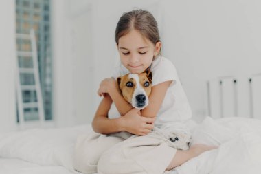 Kapalı alanda çekilen mutlu küçük çocuk en sevdiği evcil hayvanı kucaklıyor, pijama giyiyor, köpeğine sevgisini ifade ediyor, uyandıktan sonra hayvanla oynuyor, yumuşak yatakta poz veriyor. Çocuklar ve evcil hayvanlar kavramı
