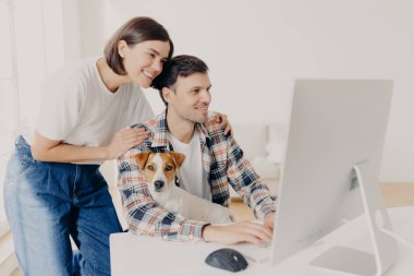 Kadınların, kocalarının omuzlarına nazikçe dokunmaları, monitörlere olumlu bakmaları, internette sörf yapmaları, yeni evleri için internetten mobilya almaları. Mutlu adam köpekle modern cihazda çalışıyor.