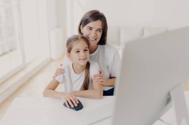 Sevecen anne küçük kızını kucaklıyor, modern bilgisayarda birlikte çalışıyor, ders için sunum hazırlıyor, zevkle monitöre bakıyor, evde boş zaman geçiriyor, internet sayfasında sörf yapıyor..