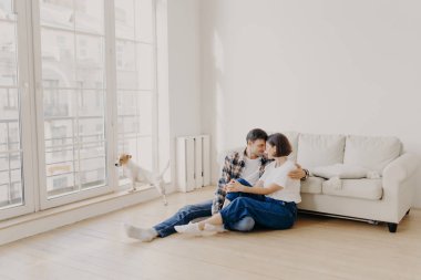 Gündelik giysiler giymiş, kucaklaşan ve birbirlerine sevgilerini ifade eden, modern apartmandaki kanepenin yanında poz veren, evcil hayvanları büyük pencereden bakan mutlu ve sevecen aile çiftlerinin yatay görüntüsü.
