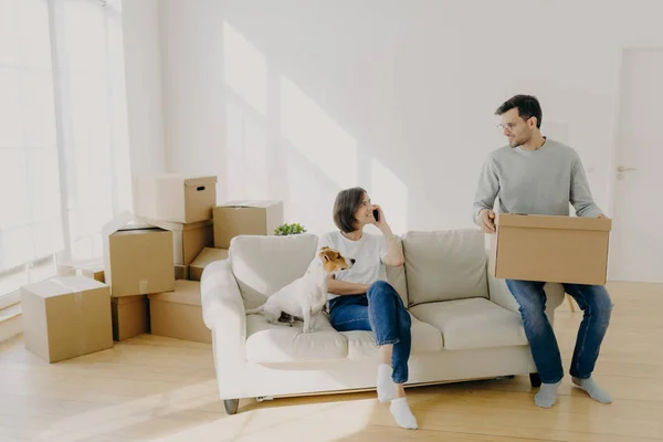 Photo of happy couple move in new home, pose on sofa with pet and boxes, have relocation, unpack boxes, female calls delivery service, carry belongings in carton containers. Welcome to new apartment