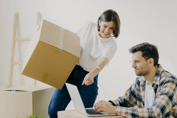 Indoor shot of happy family couple look attentively at laptop computer, search good moving company, carry personal belongings in big cardboard boxes, woman points into display of modern gadget