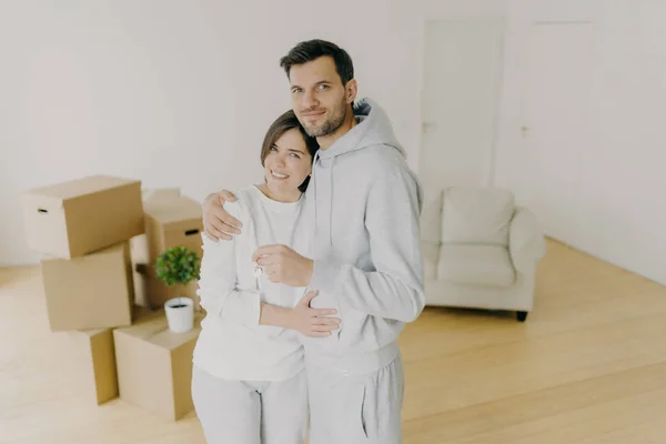 Millennial couple with plesant smiles, hug and hold key from own apartment, buy their first home, moved in shared house, stand in empty room with carton boxes in background. Ownership concept