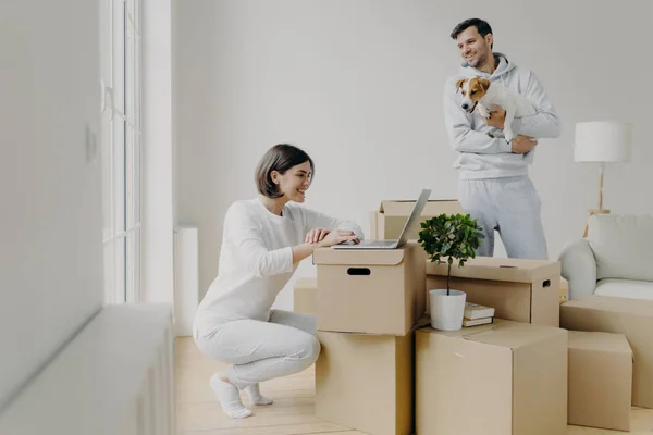Foto av lycklig kvinna arbetar på distans på bärbar dator, har glada leende, glad man i casual kläder leker med husdjur, tillbringa fritid i sitt nya hem, omgiven av kartonger i mitten — Stockfoto