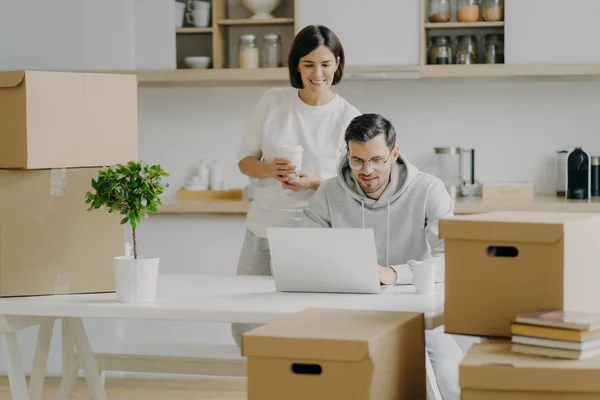 Foto de la joven alegre está detrás de su marido que trabaja en el ordenador portátil, posar en la cocina moderna de su nuevo apartamento, rodeado de cajas de cartón, pensar en el diseño moderno — Foto de Stock