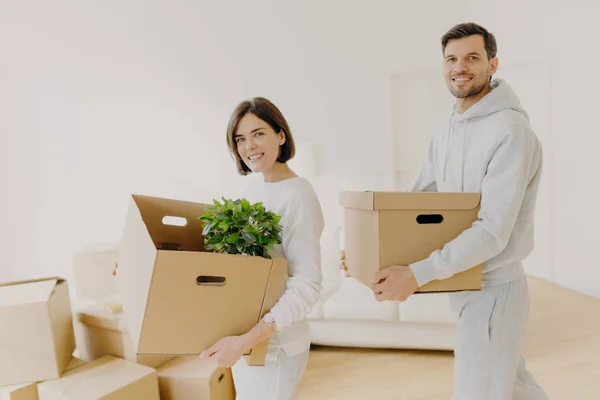 Buying real estate and mortgage loan concept. Glad woman and man carry cardboard boxes move into new home, busy with unpacking. Family property owners hold carton containers with personal stuff