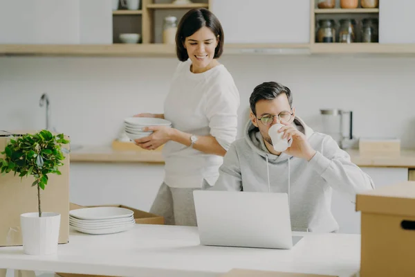忙碌的丈夫在笔记本电脑上工作，喝咖啡，戴着眼镜，穿着休闲装，妻子忙着收拾东西，端着碗碟，在搬家的日子里摆姿势在厨房里。 家庭和抵押 — 图库照片