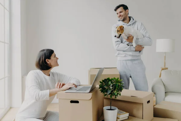 Gelukkig familie paar verhuist naar een nieuwe flat voor het leven. Vrouw werkt op laptop computer, zoekt informatie op internet, maakt online winkelen, man staat met hond, pakt spullen uit dozen. — Stockfoto