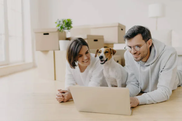 Vrolijke man en vrouw kijken film online op laptop, rusten op de vloer, ontspannen en praten, hun huisdier poseert tussen, verhuizen naar een nieuw huis, poseren in ruime woonkamer met uitgepakte dozen — Stockfoto