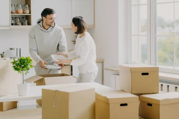 Foto de pareja familiar ocupada desempaquetar cosas personales de cajas de cartón, vestidas con ropa casual, sostener platos blancos, posar en una amplia cocina con muebles modernos, rodeado de montón de paquetes — Foto de Stock