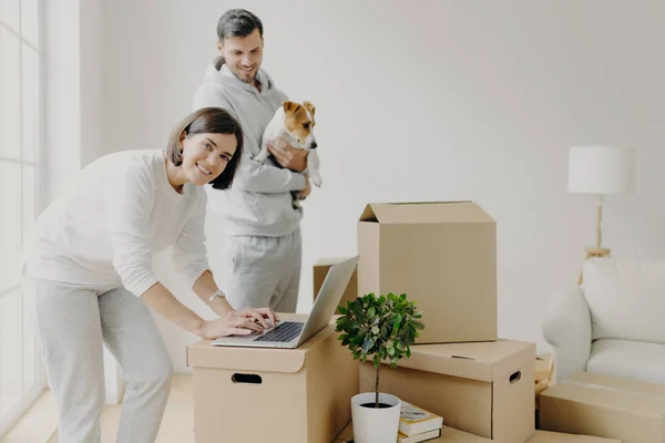 Happy home buyers pose near unpacked boxes, enjoy relocation in new house, woman searches ideas for redecoration bedroom, man carries pedigree dog, decide to do repairing of flat. Mortgage concept