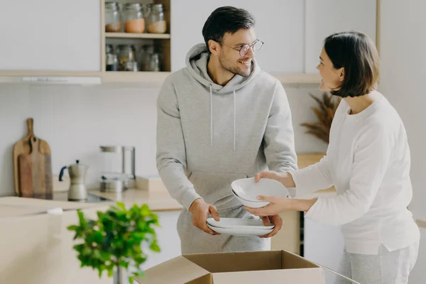 Encantada ama de casa y su marido desempaquetan cajas de cartón con vajilla nueva, llevan platos, se mudan a un nuevo apartamento, se miran con gusto, posan en el interior de la cocina agaísta. Concepto de reubicación — Foto de Stock