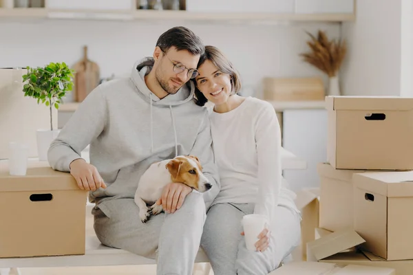 Tired married family couple drink aromatic takeaway coffee, sit closely to each other, pose with pedigree dog near big carton boxes, pose against kitchen interior, move into new modern apartment — Stock Photo, Image