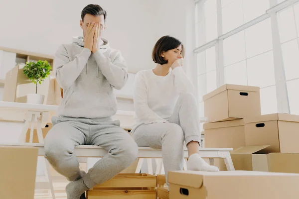Worried couple pose near stack of cardboard boxes, have to move in other flat, have no money for rent, have dejected sad expressions, sit together in white room, have family conflict and divorce — Stock Photo, Image