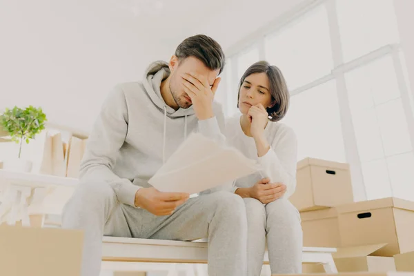 Distressed family couple have debt problem, being not able to pay apartment rent, have to leave house, man holds papers from bank, manage domestic budget, wife calms husband, pose in empty room