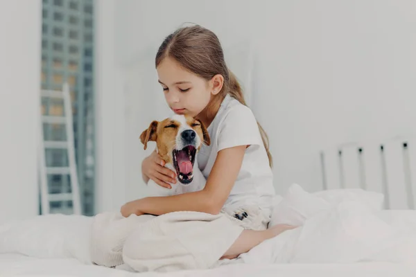 Bella dai capelli piccola ragazza con coda di cavallo indossa pigiami casual, abbraccia cane che sbadiglia, posare insieme su un letto confortevole, esprime amore e cura per gli animali domestici, trascorre il fine settimana mattina a casa — Foto Stock