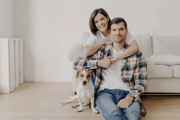 Mutlu esmer kadın kocasını sevgiyle kucaklıyor, iyi bir ruh hali içinde, olumlu gülümsüyor. Karı, koca ve köpek, yeni konutun oturma odasında birlikte poz verirler, rahatlığın tadını çıkarırlar. Birbirlerine aşık çiftler.