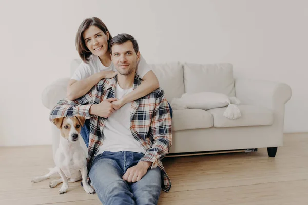 快乐女人的照片拥抱她的丈夫 在沙发上摆姿势 小狗坐在靠近主人的地板上 带着快乐的表情看着镜头 有着浪漫的关系 全家福 人与家庭 — 图库照片