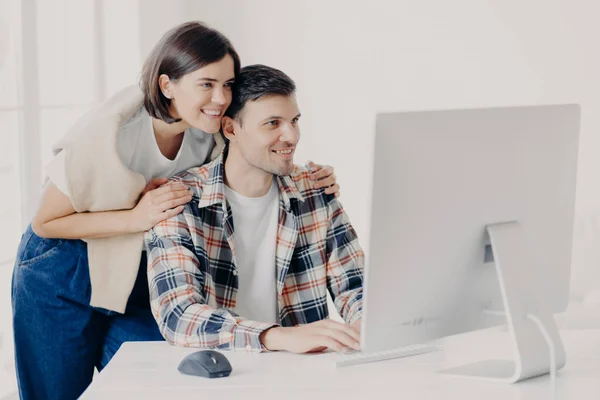 年轻夫妇合作开发网站 共同开发互联网项目 亲热的妻子在支持下拥抱丈夫 专注于监控 键盘信息 与互联网相连 — 图库照片