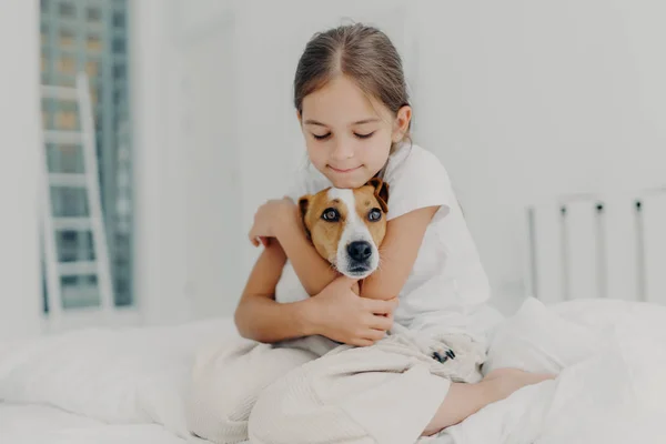 Lo sparo dell'interno di bambino piccolo felice abbraccia l'animale domestico preferito, vestito in pigiama, esprime l'amore al suo cane, gioca con animale dopo essersi svegliato, posa su letto morbido. Concetto di bambini e animali domestici — Foto Stock