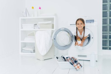 Çekici görünümlü küçük bir kız, eğleniyor ve çamaşır makinesinin içinde poz veriyor, deterjan tutuyor, çamaşır yıkamaya hazırlanıyor, çamaşır makinesine çamaşır koymaya hazırlanıyor. Evde çamaşır günü