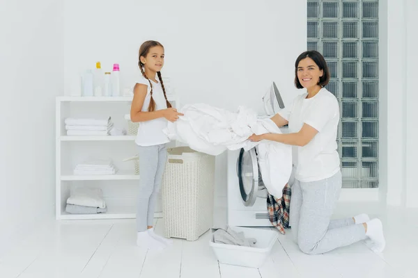 Ainda bem que a dona de casa lava-se com um ajudante adorável. Mãe e filha lavar roupas na lavanderia, roupa de cama de carga na máquina de lavar. Mulher fica de joelhos perto da máquina de lavar roupa. Conceito de trabalho doméstico — Fotografia de Stock