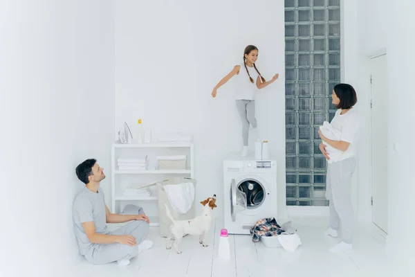 La familia feliz hace la colada en casa, el padre se sienta en el suelo en la pose de loto, la madre se para con la toalla blanca, mira al niño que baila felizmente en la lavadora, el perro pedigrí cerca. Funciones domésticas . — Foto de Stock