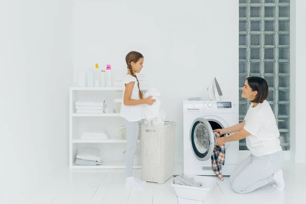 Mulher morena ocupada carrega máquina de lavar roupa com roupas sujas, sua filhinha ajuda, fica perto do cesto e lava roupas. Mãe e criança felizes na sala de lavagem. Conceito de trabalho doméstico . — Fotografia de Stock