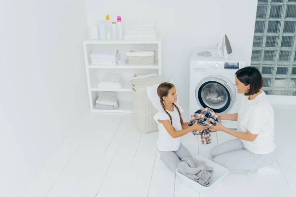 Little girl helper and her mother pose in laundry room near washing machine, sit on white floor, hold dirty shirt, do washing at home during weekend, load clothes. Family chores and domestic work