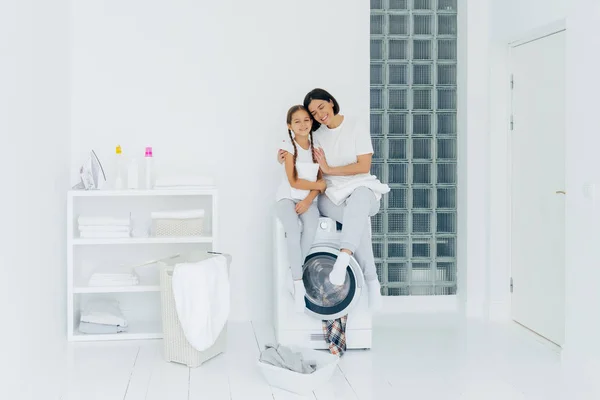 Feliz madre e hija se divierten, abrazan y expresan amor entre sí, se visten con ropa casual, se sientan en la lavadora, se preparan para el lavado, estar en el amplio lavadero blanco, lavabo con ropa sucia — Foto de Stock