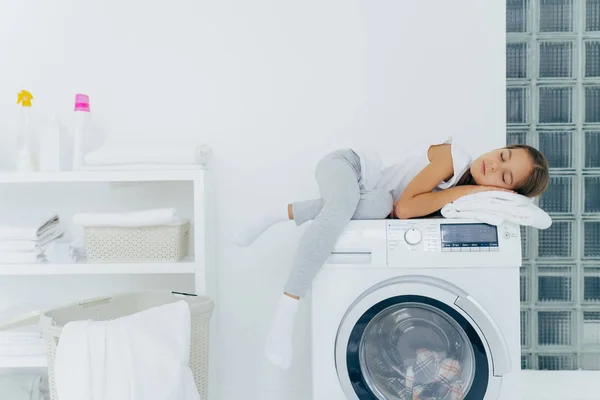 Tiro horizontal de menina tem descanso na máquina de lavar roupa, se sente cansado depois de ajudar a mãe sobre a casa, dorme em poses lavadora na lavanderia com console, detergentes, roupa de cama ao redor. Rotinas domésticas — Fotografia de Stock