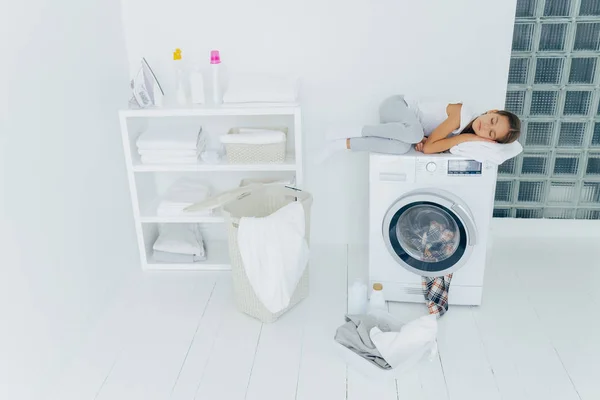 Tiro de criança pequena cansada dorme na máquina de lavar roupa, usa toalha macia branca como travesseiro, tem sonhos agradáveis, cesta e bacia no chão branco, sente fadiga depois de carregar roupas na máquina de lavar roupa, estar em casa — Fotografia de Stock