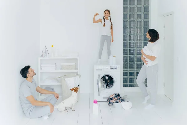 Chica alegre fuerte levanta el brazo, muestra bíceps, divierte a los padres, tienen descanso después de lavar la ropa, posar en un amplio lavadero blanco con mascotas, lavadora y agentes de limpieza. Familia y tareas domésticas — Foto de Stock