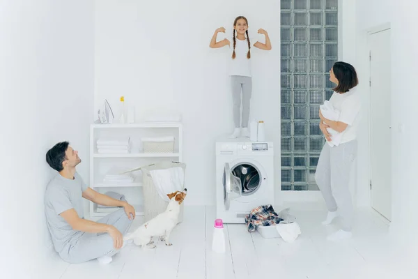 Foto de colegiala feliz posa encima de la lavadora, muestra los músculos, levanta los brazos, listo para ayudar a los padres con el lavado o la ropa. Mujer y hombre posan en la sala de lavado con perro y niño pequeño — Foto de Stock