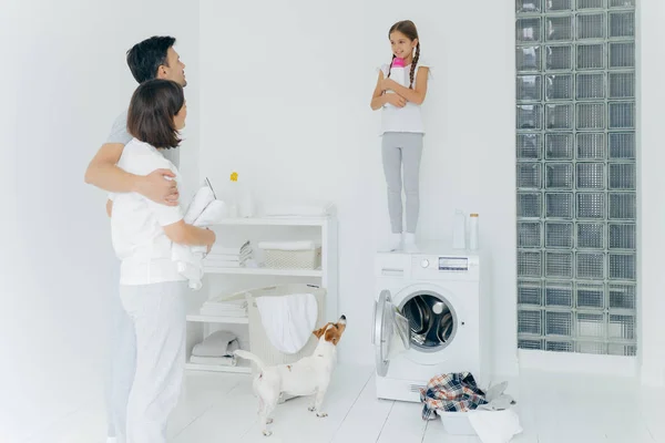 La familia feliz lavan juntos en el lavadero. Padre y madre se abrazan mientras miran a la hija en la parte superior de la lavadora, rodeada de lavabo de ropa para lavar, mascotas y armario con ropa plegada — Foto de Stock